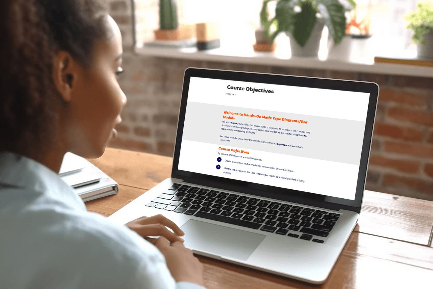 A woman sits at a wooden desk, focused on a laptop screen displaying course objectives. Sunlight illuminates the room, highlighting a few potted plants on the windowsill, creating a calm, focused atmosphere.