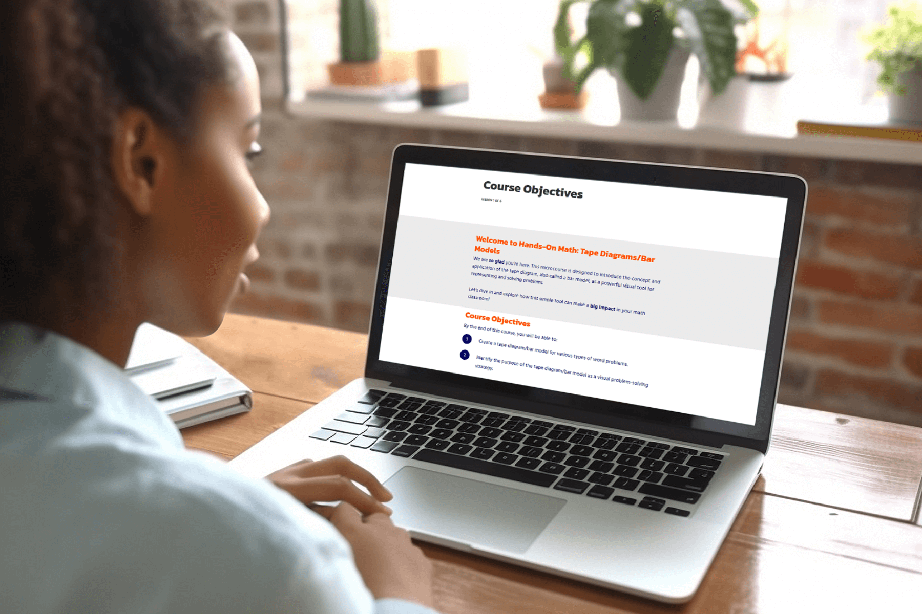 A woman sits at a wooden desk, focused on a laptop screen displaying course objectives. Sunlight illuminates the room, highlighting a few potted plants on the windowsill, creating a calm, focused atmosphere.