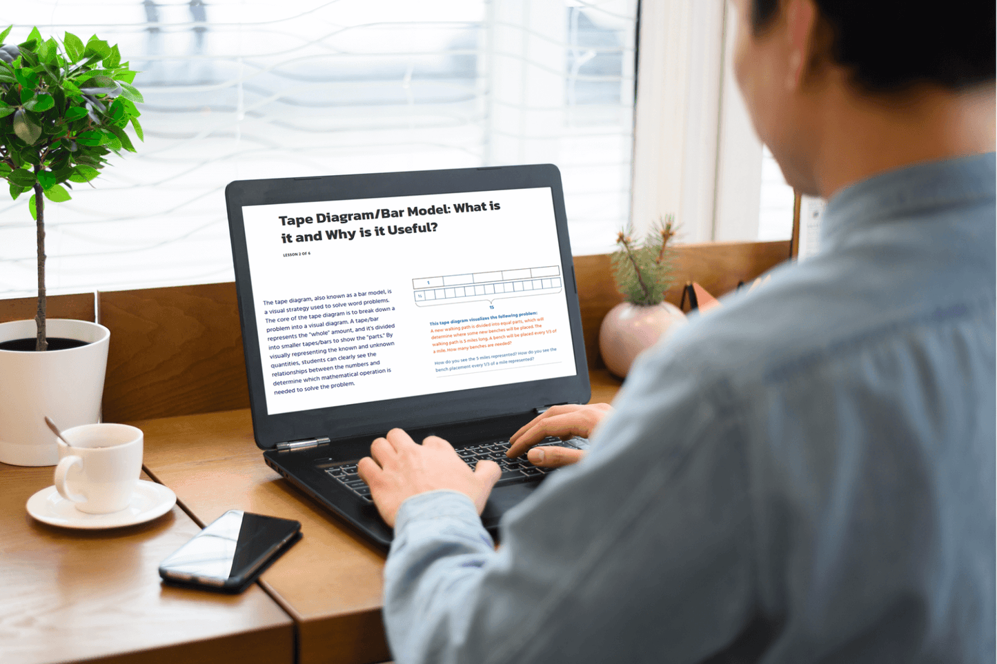 A person in a casual blue shirt works on a laptop at a wooden desk with a plant, phone, and coffee cup. The screen displays a tape diagram article.