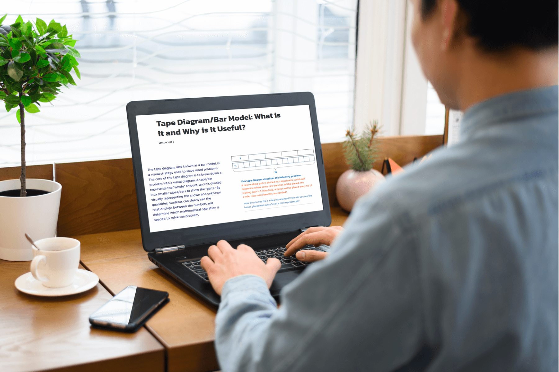 A person in a casual blue shirt works on a laptop at a wooden desk with a plant, phone, and coffee cup. The screen displays a tape diagram article.
