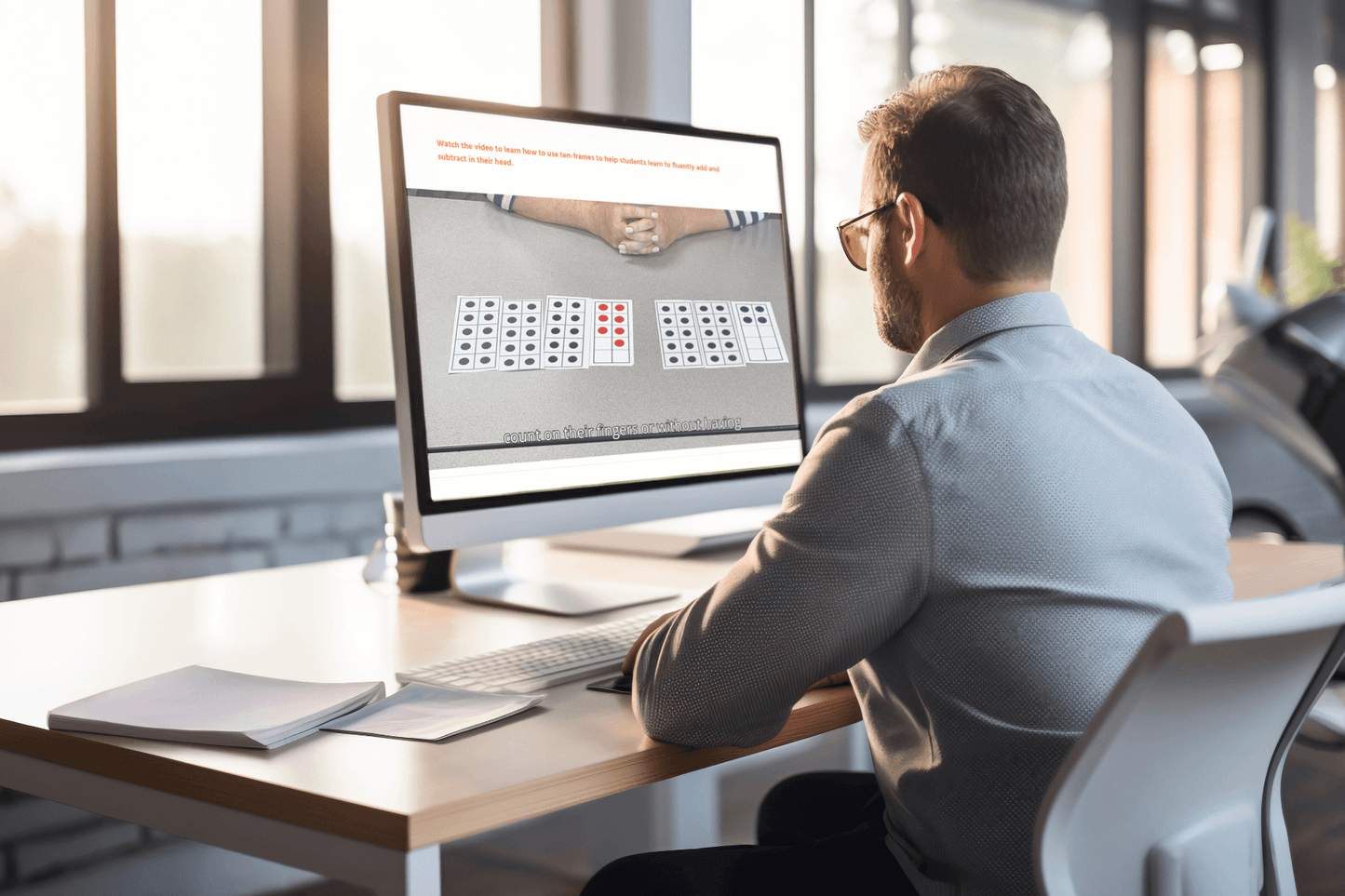 A man sits at a desk in a modern office, focused on a computer screen displaying a video how use Ten-Frames. The sunlight filters through large windows.