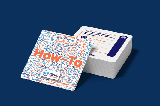 A deck of cards lies on a blue surface. The top card is visible, featuring a colorful word cloud with large "How-To" text. Keywords include "brave," "unstuck," "hearts," and "minds."