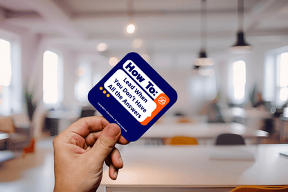 A hand holds a square card with the text "How To: Lead When You Don’t Have All the Answers" in a modern office, suggesting guidance in leadership.