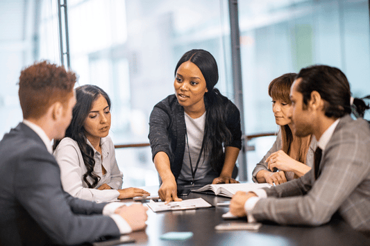 Person standing and pointing to a document on the table while leading a discussion with four seated colleagues in a conference room.