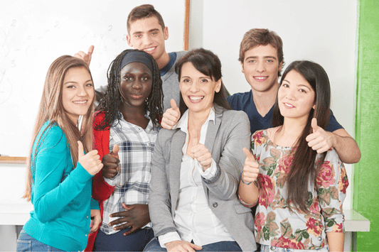 A group of six people, including one adult and five teenagers, stand close together indoors, smiling and giving thumbs-up gestures toward the camera. They appear to be in a cheerful, encouraging setting, possibly a classroom or school environment.