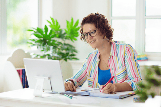 An adult sits at a desk, smiling while working on a laptop. They hold a red pen and write in a notebook, with open books and papers spread out. A bright window and green plants are in the background.
