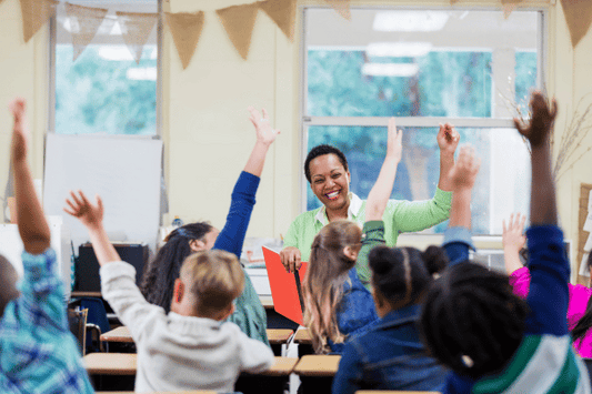 A smiling teacher stands at the front of a classroom holding a folder while many students eagerly raise their hands to participate. The classroom is bright with natural light coming through large windows.