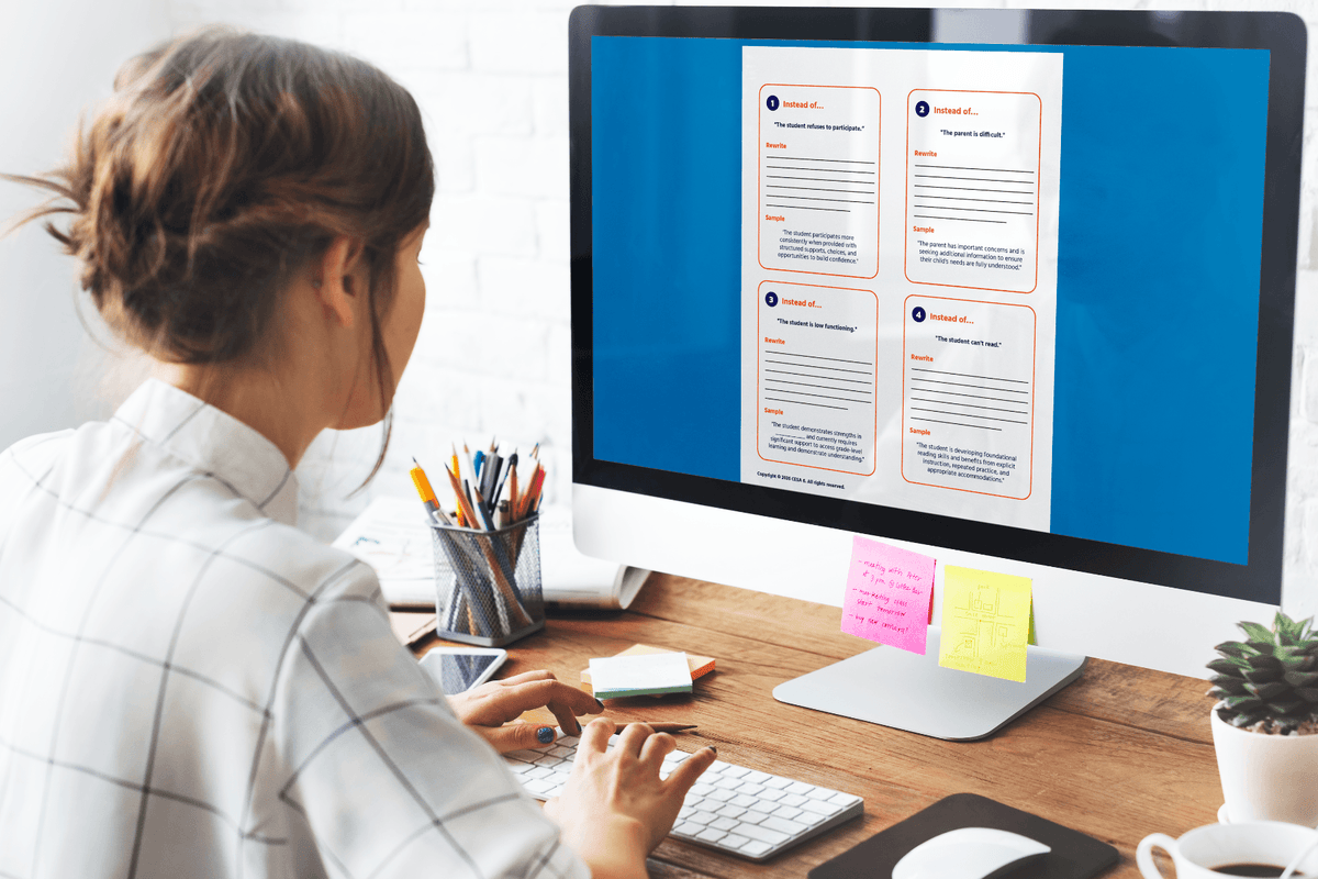 Woman working at a desktop computer displaying "Strengths-Based Language Makeover Cards" online resource, with a keyboard, stationery, sticky notes, and a plant nearby.