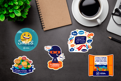 Five colorful stickers are displayed on a desk, each with a unique design promoting multilingualism and cultural connection. The designs feature messages such as “Everyone Smiles in the Same Language,” “Being Bilingual is My Superpower,” “Knowledge of Languages is the Doorway to Wisdom,” “To Have Another Language is to Possess a Second Soul,” and “To Learn a Language is to Have One More Window from Which to Look at the World.”