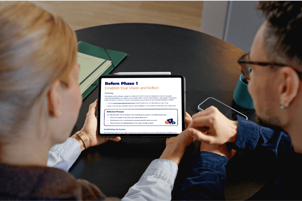 Two people seated at a round table looking at a tablet screen showing a digital document or webpage. One person holds the tablet.