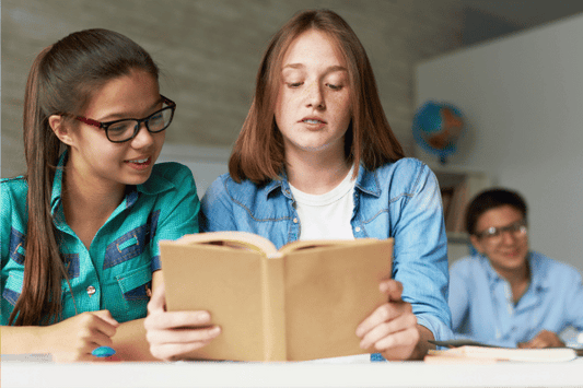 Two teenage students are sitting side by side at a desk, reading a book together. One student, wearing glasses and a teal plaid shirt, is smiling and looking at the book, while the other student in a denim shirt reads aloud.