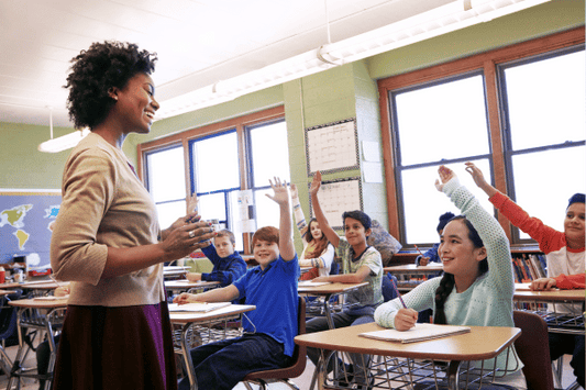An educator stands at the front of a middle-grade classroom, speaking and smiling while several students seated at desks eagerly raise their hands. 