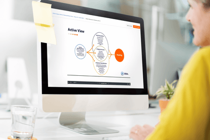 A person in a yellow shirt views a self-paced learning course on an iMac. The screen shows a diagram titled "Active View." A sticky note is attached to the monitor.