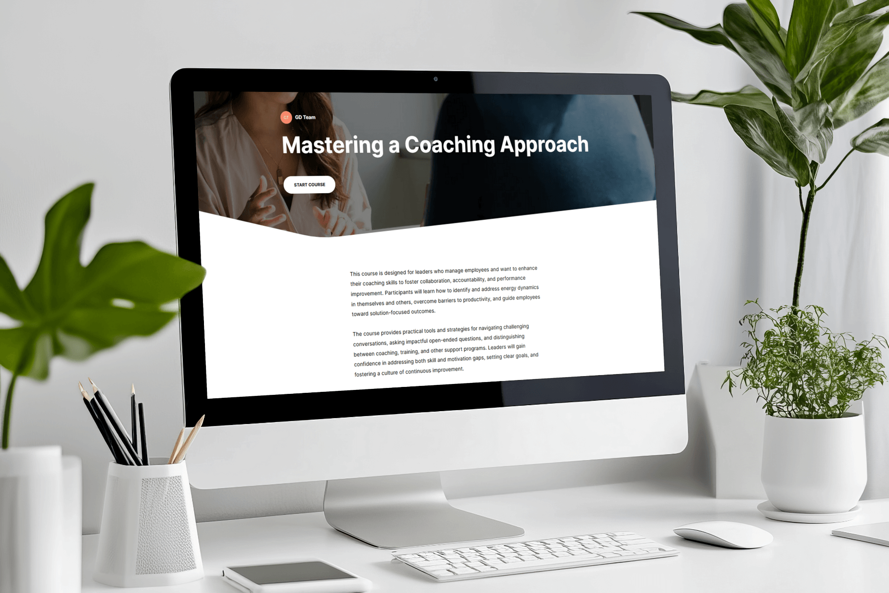 A computer screen displaying "Mastering a Coaching Approach" on a webpage with a start button. The desk features green plants and stationery, creating a fresh, professional atmosphere.