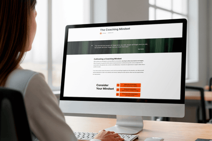 A person sits at a desk, using a computer displaying an article titled "The Coaching Mindset." The screen shows text and an orange section, creating a focused, professional tone.