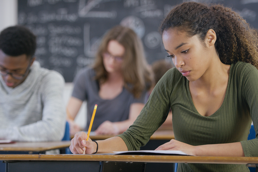 A diverse group of high school students seated at desks in a classroom, focused on taking a test. A young student in the foreground, wearing a green long-sleeve shirt, is writing with a pencil. A chalkboard filled with mathematical equations is visible in the background.