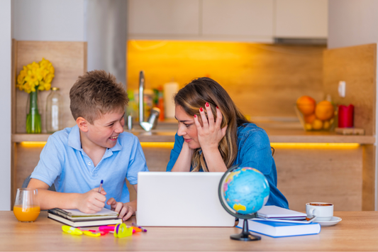 A mother and her young son sitting at a kitchen table engaged in tutoring. The boy, wearing a light blue polo shirt, is smiling while writing in a notebook. 