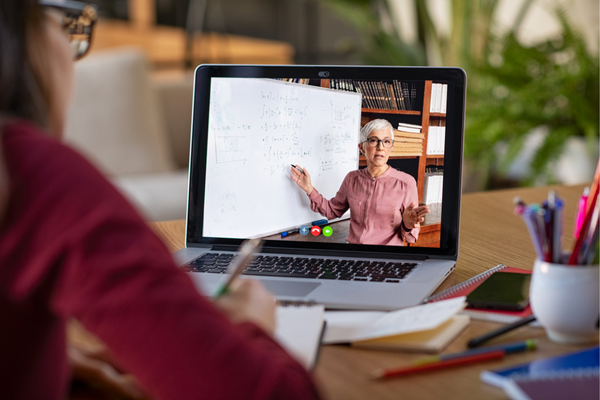 A student wearing a red sweater sits at a desk, taking notes while attending an online lesson on a laptop. The screen displays a teacher explaining a math concept using a whiteboard, with bookshelves in the background. The desk is cluttered with notebooks, pencils, and other stationery.