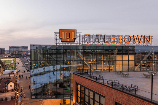 An exterior view of the TitletownTech building shows a modern glass and brick structure with a large illuminated “TITLETOWN” sign on top. The reflection of an ice skating rink and string lights is visible in the building’s glass facade.