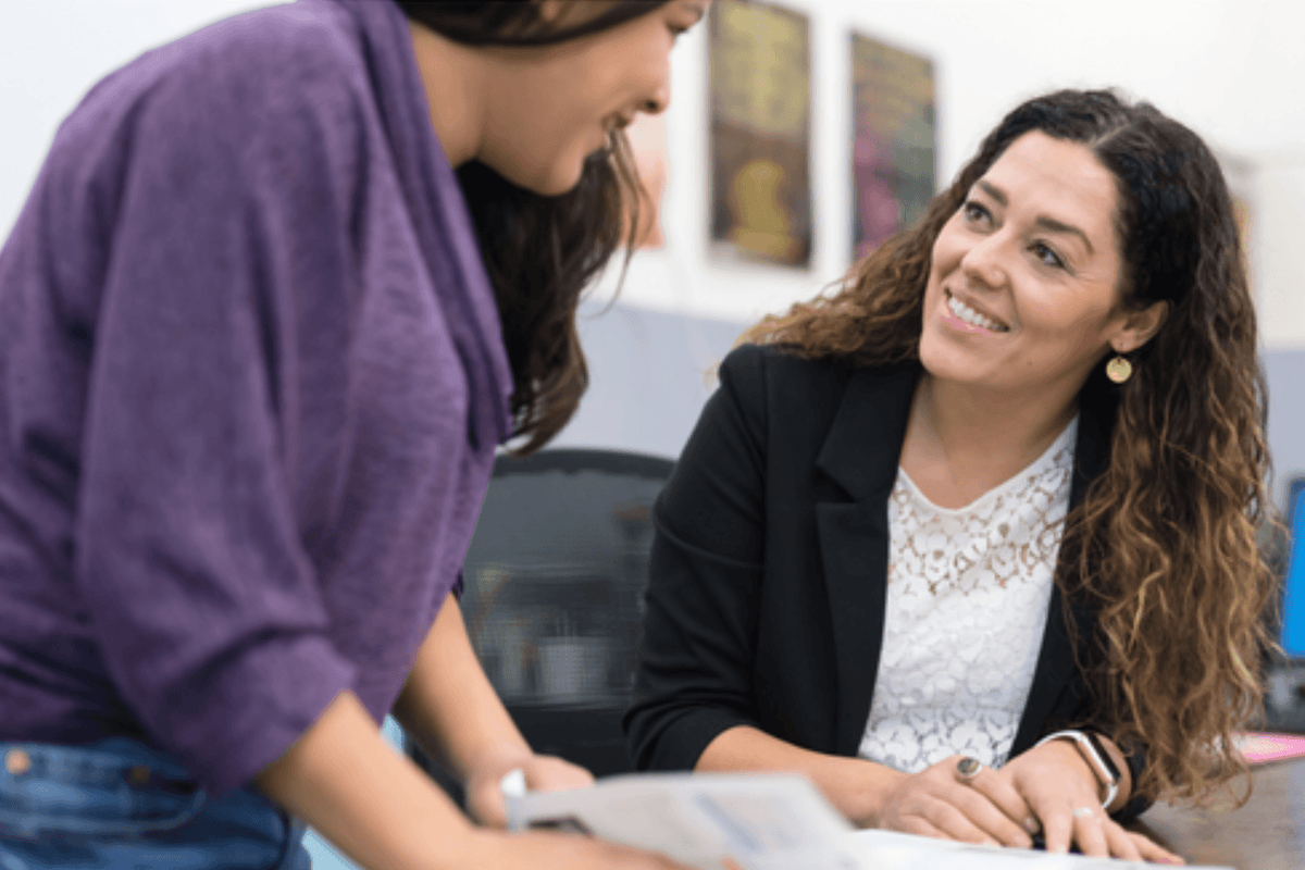 Two people collaborate at a desk, reviewing documents; one smiles warmly at the other in a bright office, conveying teamwork and support.