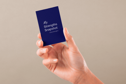 A hand holding a blue card that reads "My Strengths Snapshot, CliftonStrengths." The background is a neutral beige, conveying a calm and focused tone.
