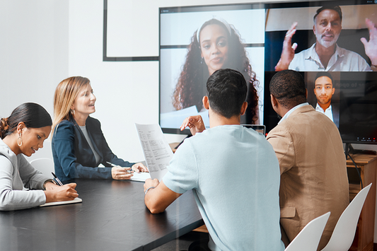 A group of five professionals sit around a modern conference table, participating in a hybrid meeting. Four individuals are shown on a large screen at the front of the room, joining the meeting virtually. The in-person attendees are taking notes, reviewing papers, and engaging with the virtual participants. The setting is a bright office space with white walls and minimal decor.