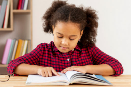 Young girl sitting at table, using finger to point while reading lines from a book.