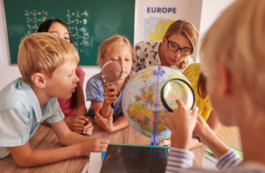 A group of elementary-aged children is gathered around a table with a globe in the center. One child is holding a magnifying glass up to the globe, while others look on with curiosity. An adult, likely a teacher, leans in closely, engaging with the students. A chalkboard with math problems and a map labeled “Europe” are visible in the background, indicating a classroom setting.