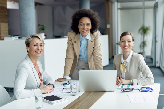 Three professional women smiling and working together in an office setting. One woman is standing and leaning on the table, while two are seated with a laptop, documents, and glasses of water on the table.