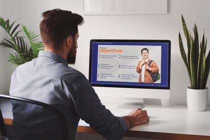 A person sits at a desk viewing an online learning module titled “IEPs and 504 Plans: A Guide for Educators” on a desktop computer. The slide on the screen is titled “Learning Objectives” and includes a list of six goals related to understanding IEPs and 504 Plans. On the right side of the slide is a photo of a smiling student wearing glasses and a backpack. The workspace features a potted plant and is brightly lit with natural light from a nearby window.