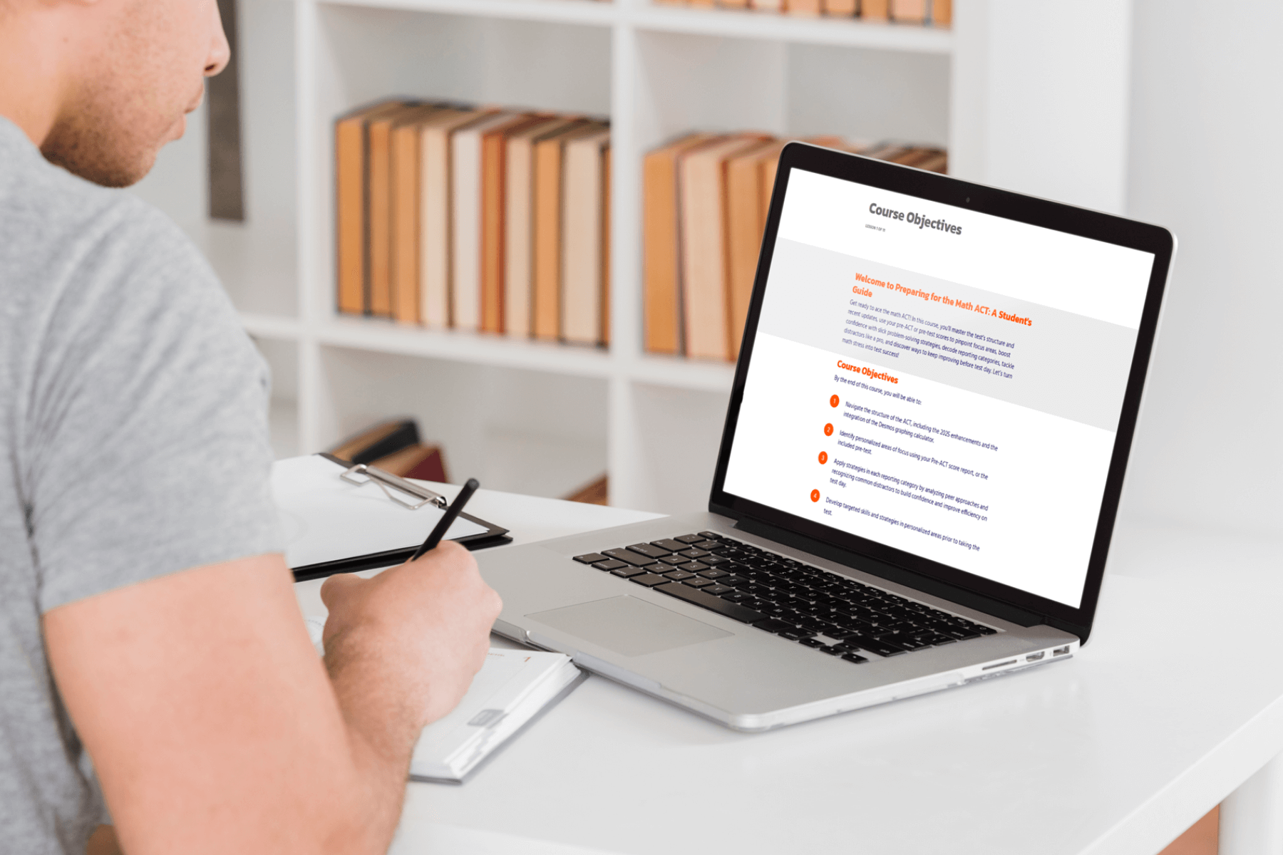 A person in a gray shirt is taking notes while viewing a laptop displaying course objectives. The setting is a bright room with a white desk and bookshelves.