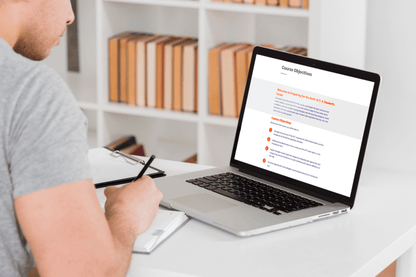 A person in a gray shirt is taking notes while viewing a laptop displaying course objectives. The setting is a bright room with a white desk and bookshelves.