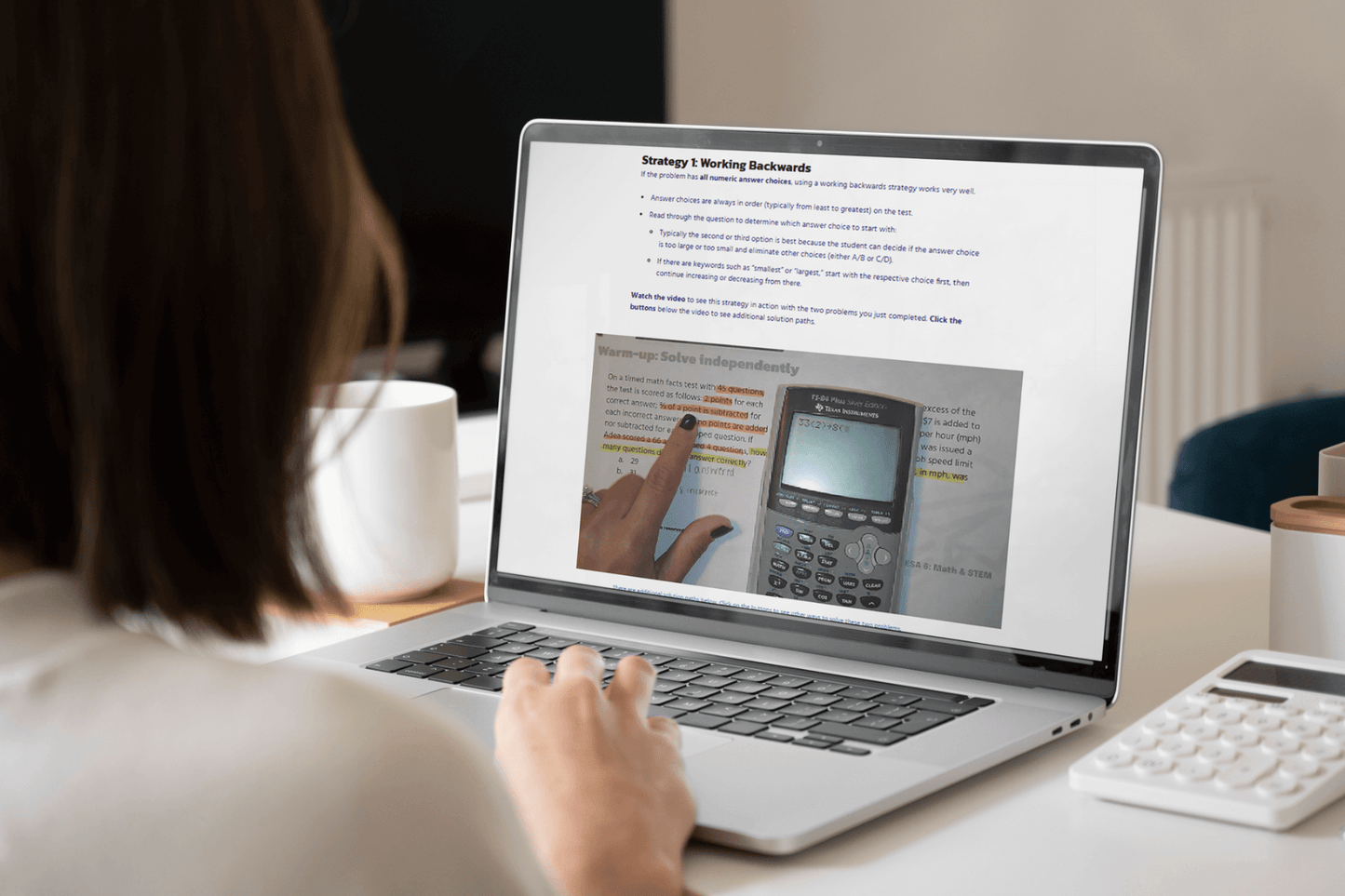 A person views educational content on a laptop screen, featuring problem-solving strategies and a calculator image. The setting is a bright workspace.