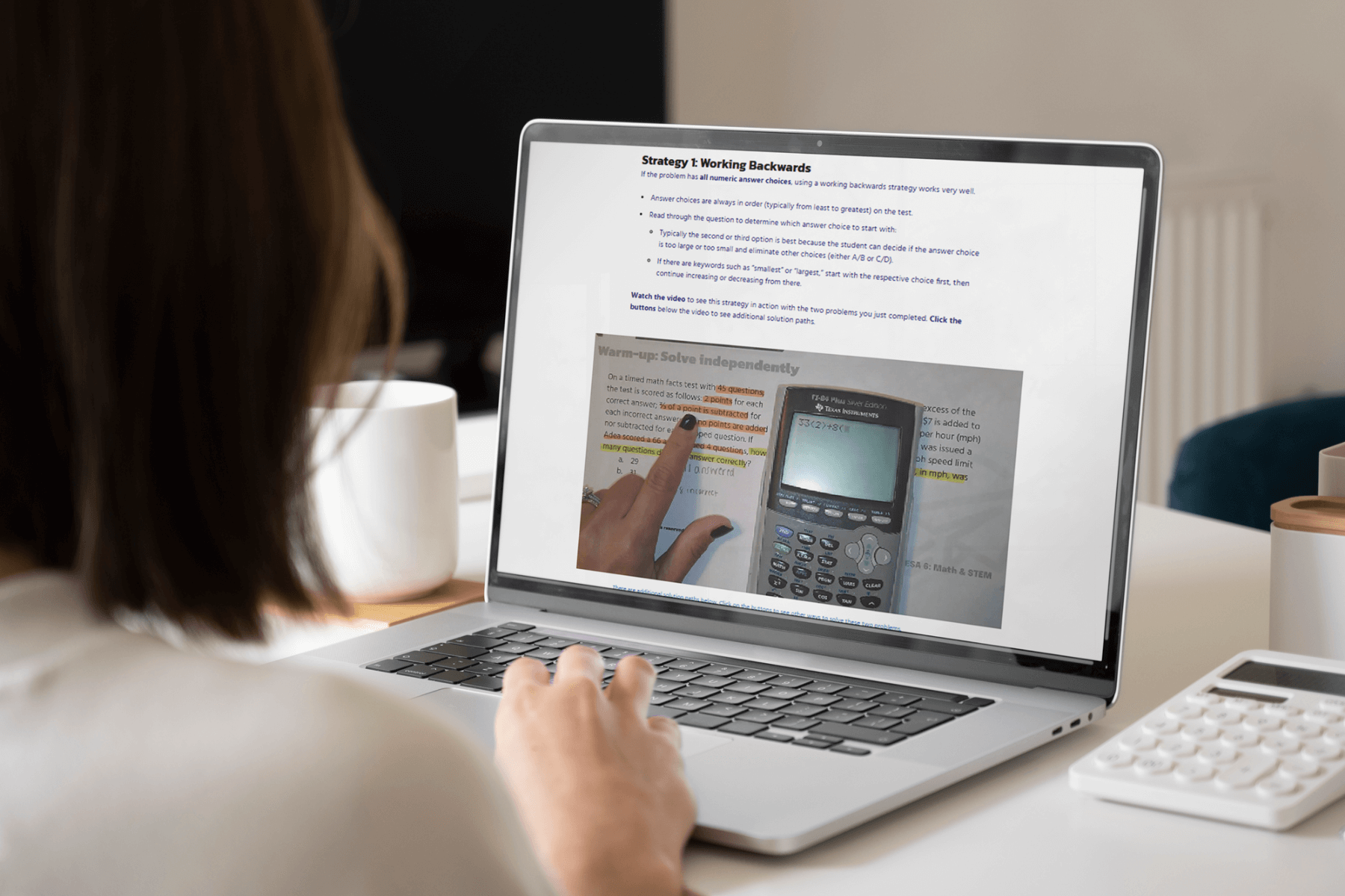 A person views educational content on a laptop screen, featuring problem-solving strategies and a calculator image. The setting is a bright workspace.
