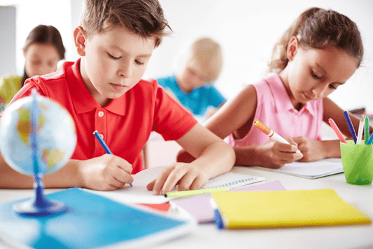 Children sit at classroom desks, focused on writing in their notebooks. A small globe and colorful folders are on the table. The students appear engaged in their work, suggesting a structured classroom activity.