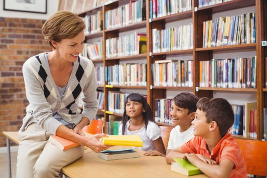 An adult sits at a table in a library, smiling and handing books to three seated children. Bookshelves filled with colorful books line the background. The children are attentively looking at the adult, appearing engaged and cheerful.