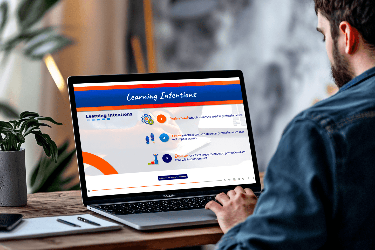 A man sits at a desk looking at a laptop displaying a presentation titled "Learning Intentions," focusing on professionalism and personal development.
