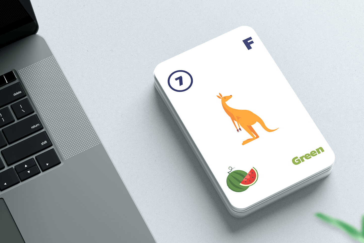 A stack of randomized grouping cards placed on a light gray desk next to a laptop. The top card features a yellow kangaroo illustration, the number 7, the letter 'F,' a watermelon image, and the word 'Green' in green text.