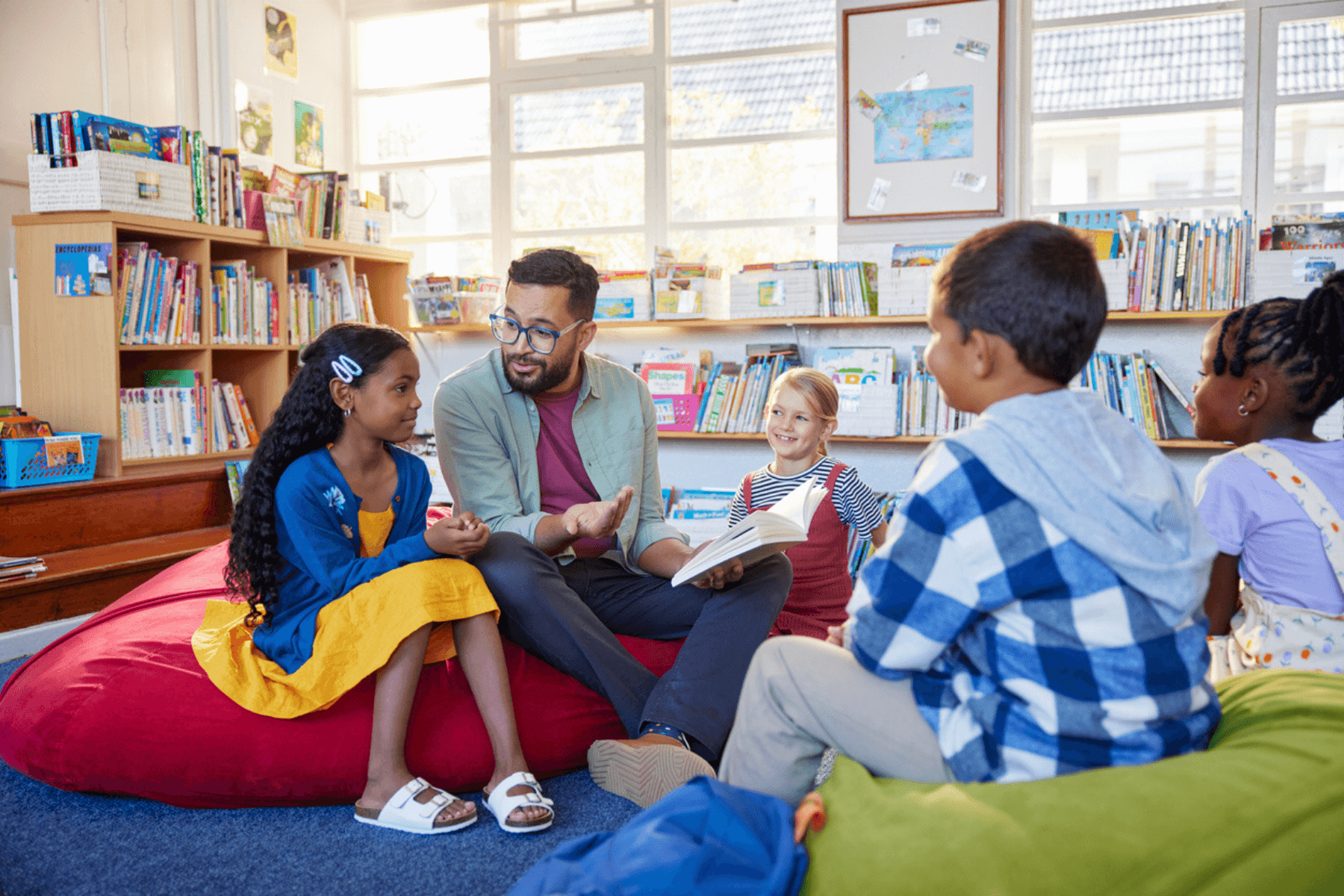 A teacher is sitting on a beanbag chair in a well-lit classroom library, engaged in conversation with a group of young students seated around him on beanbags and cushions. Bookshelves filled with children's books line the walls, and large windows let in natural light. The atmosphere appears relaxed and inviting, with students smiling and interacting attentively.