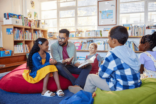 A teacher is sitting on a beanbag chair in a well-lit classroom library, engaged in conversation with a group of young students seated around him on beanbags and cushions. Bookshelves filled with children's books line the walls, and large windows let in natural light. The atmosphere appears relaxed and inviting, with students smiling and interacting attentively.