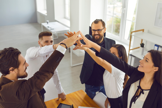 A group of professionally dressed adults stands around a conference table in a bright office, reaching into the center for a group high-five. They are smiling and appear to be celebrating or collaborating successfully, with natural light streaming in from large windows.