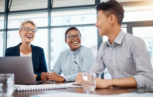 Three adults are seated around a table in a bright, modern office space, smiling and engaged in conversation. A laptop, notebooks, and a glass of water are on the table in front of them, suggesting a collaborative meeting or discussion.