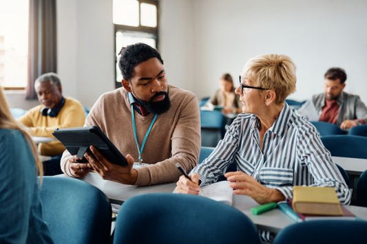 Two adults sit in a classroom, engaged in conversation. One person is holding a tablet, while the other is writing in a notebook. Additional people are seated in the background, suggesting a workshop or collaborative training setting.
