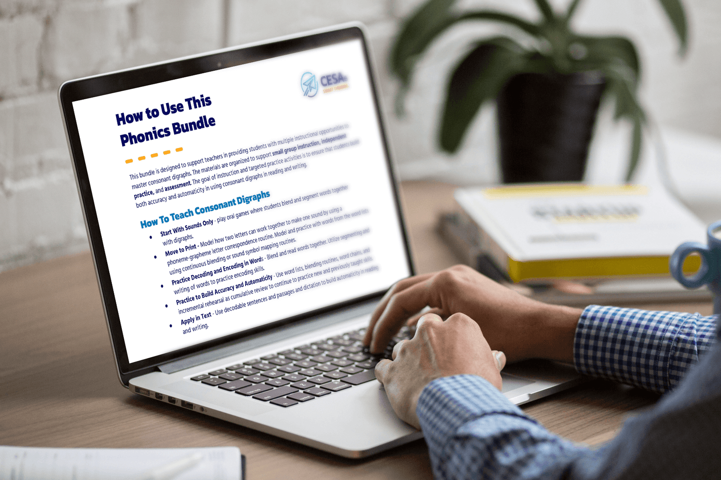 A person in a checked shirt is using a laptop displaying a document titled "How to Use This Phonics Bundle." A plant and books are on the desk.