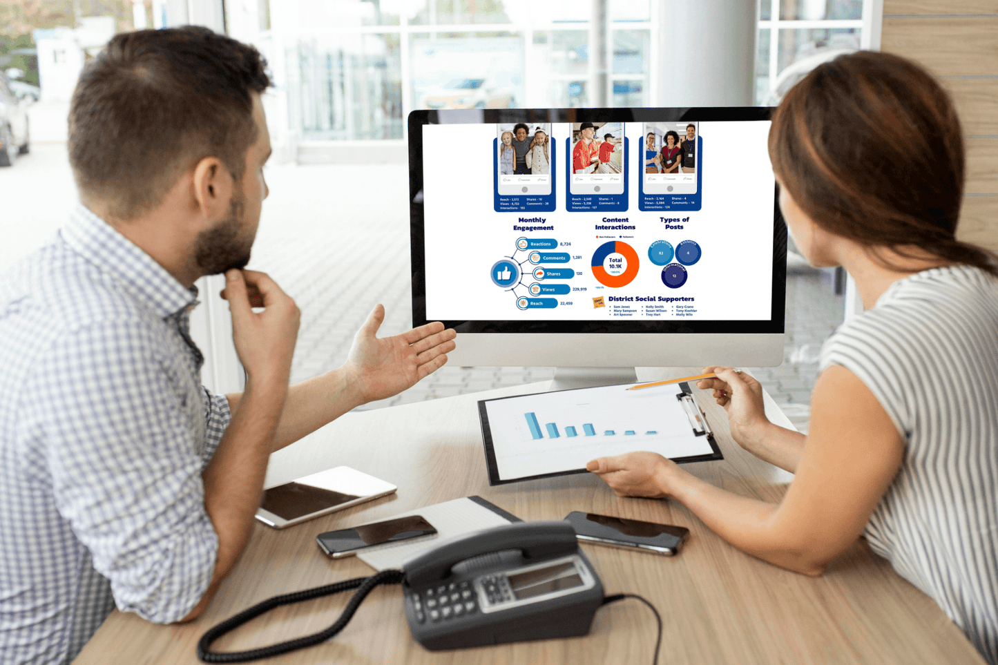 A man and woman discuss data displayed on a desktop screen showing graphs and charts. The setting is an office with a focused, collaborative atmosphere.
