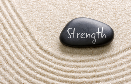 A smooth black stone rests on fine, light-colored sand that has been carefully raked into concentric curves. The word "Strength" is written in white cursive letters across the surface of the stone. The overall scene evokes calmness and focus, reminiscent of a Zen garden or mindfulness practice.