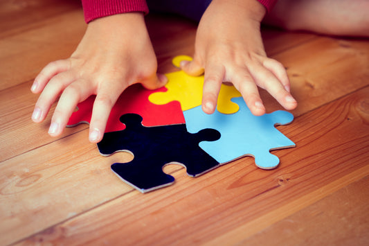 The image shows a close-up of a child's hands placing colorful puzzle pieces on a wooden floor, with the pieces in red, yellow, blue, and black.