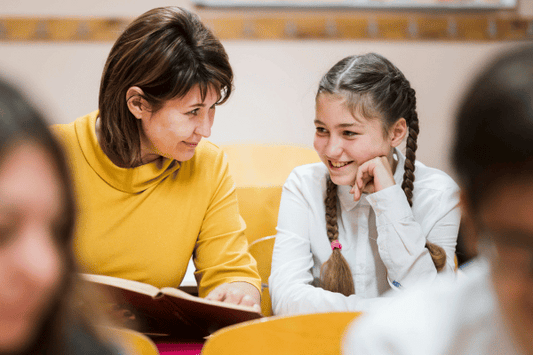 A teacher in a yellow top sits closely with a smiling young student who has braided hair, engaging in a warm and focused reading session. They are seated in a classroom, and the student looks up at the teacher with enthusiasm while resting their chin on her hand.
