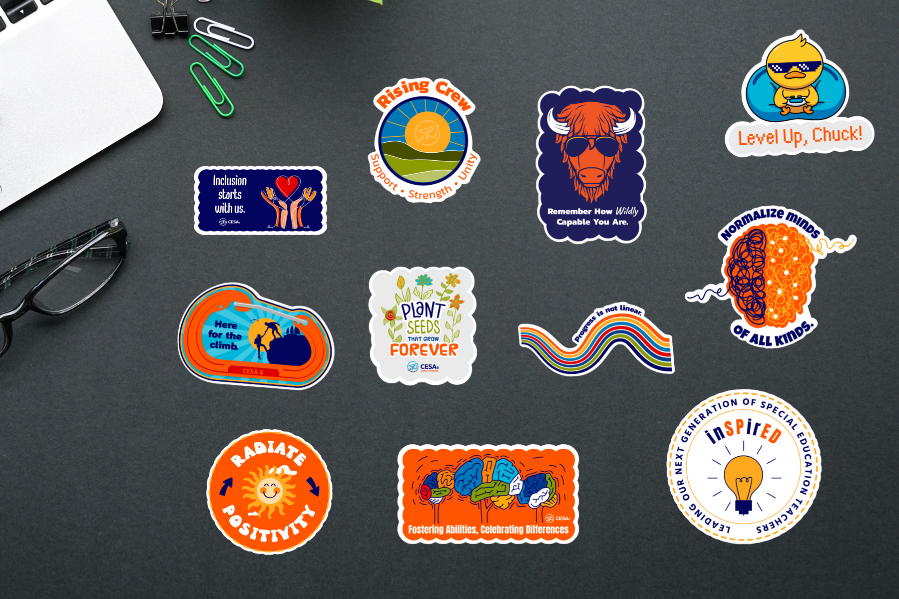 A collection of colorful stickers with motivational and inclusive messages displayed on a dark surface. The stickers include phrases like 'Inclusion starts with us,' 'Radiate Positivity,' 'Normalize minds of all kinds,' 'Fostering Abilities, Celebrating Differences,' and 'Plant seeds that grow forever.' Other designs feature a buffalo with sunglasses, a duck with a gaming controller, and a sun with a smiley face. Office supplies such as paper clips and glasses are also present in the scene.