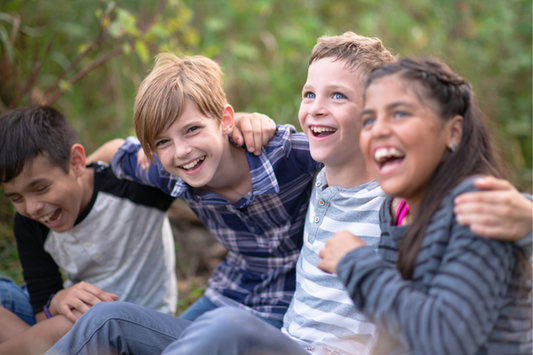 This image features four children sitting together outdoors, laughing, and having fun. They are close together, with their arms around each other.